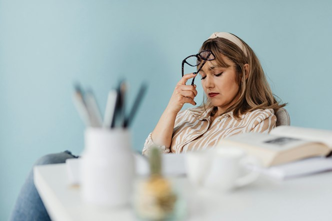 A woman sitting at a desk, holding her glasses with her eyes closed, appearing fatigued. The desk is cluttered with books, a pencil holder, and a blurred mug in the foreground. She is wearing a striped shirt and has a beige headband.