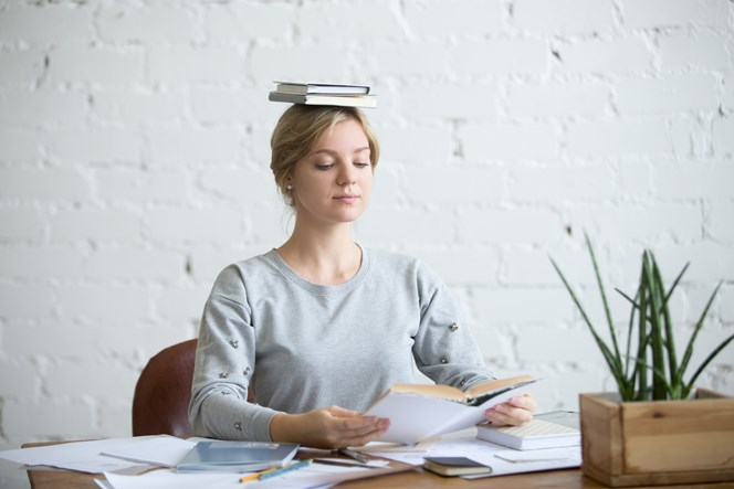 A woman sitting at a desk with books balanced on her head, reading a book, against a white brick wall background.
