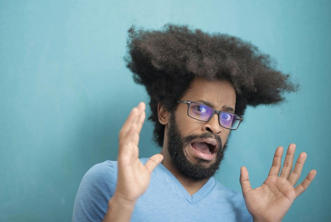 A man with an afro hairstyle and glasses looks surprised, raising his hands and eyebrows in a light blue shirt against a turquoise background.