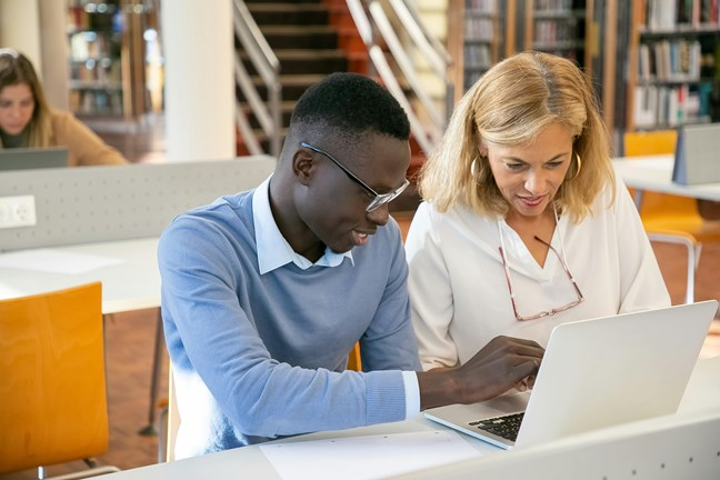 A young man and a woman collaborating on a laptop in a library setting, sitting at a white desk with bookshelves and other people in the background.