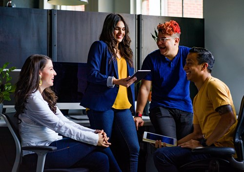 Four people having a casual meeting in an office setting, with one person holding a tablet and the others smiling and talking.