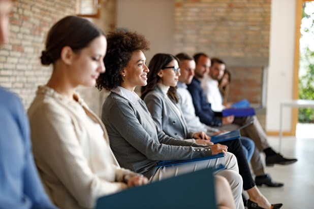 A diverse group of people sitting in a row, waiting, holding folders; they appear to be in a formal setting, possibly for an interview or meeting.