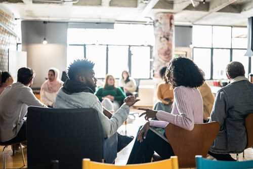 A diverse group of people sitting in a circle and engaging in conversation in a bright, modern room with large windows and a casual setting.