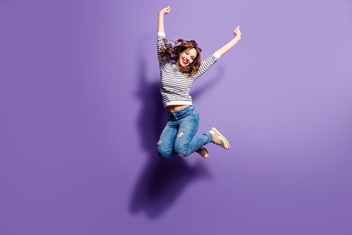 A person wearing a striped shirt and jeans is joyfully jumping against a purple background.