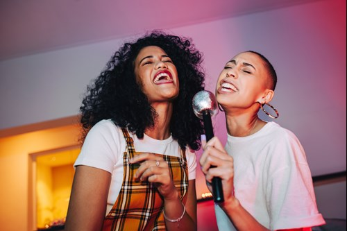 Two people joyfully singing into a microphone, with one holding it and the other wearing a plaid dress, both smiling and relishing the moment.