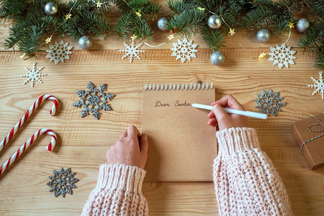 A person wearing a pink knitted sweater writes 'Dear Santa' on a notepad, surrounded by festive decorations including candy canes, decorative snowflakes, and a small gift box on a wooden table under fir tree branches adorned with ornaments.