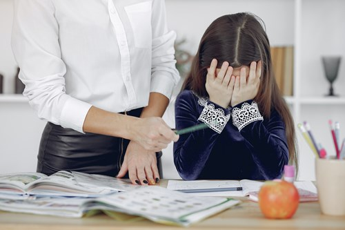 A child in a blue velvet dress covers her face with her hands, sitting at a table cluttered with books and pencils. A standing adult in a white shirt points at the child's workbook with a pen.