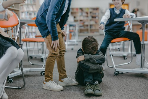 A child sits on the floor with arms wrapped around their knees, appearing distressed, while another stands nearby and a third sits in an orange chair, smiling.