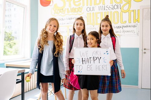 Four smiling schoolchildren standing in a classroom hold a sign reading, 'We are happy to be here.'