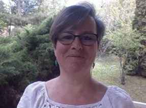 A person with short hair and glasses wearing a light-coloured top, standing outdoors with trees and greenery in the background.