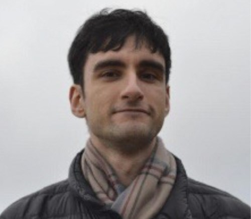 A person with short dark hair wearing a plaid scarf and a quilted jacket, set against a neutral background.