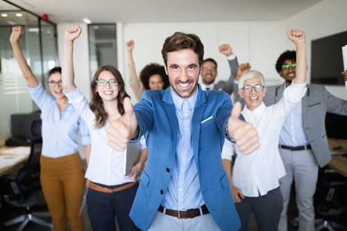 A group of diverse office workers celebrating with smiles and raised fists, with a man in a blue blazer giving thumbs up in the foreground.