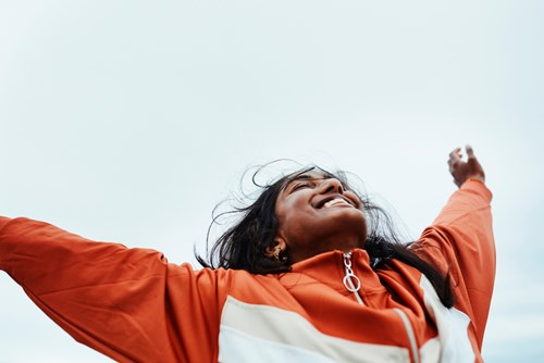 Person wearing an orange jacket joyfully stretching their arms upwards against a cloudy sky background.
