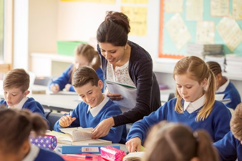 A teacher assists a student in a classroom filled with children wearing matching blue uniforms as they work on assignments at their desks.