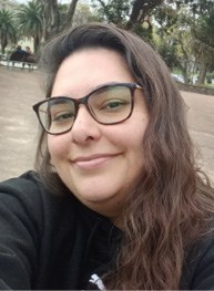 A person with long hair, wearing glasses and a dark hoodie, smiling while taking a selfie in an outdoor park setting.