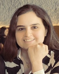 A person with long dark hair smiling at the camera, wearing a black and white patterned shirt, with a hand resting on their chin.