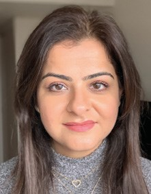 A woman with long brown hair wearing a grey turtleneck and a small heart-shaped necklace.