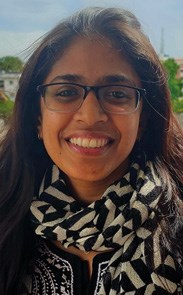 A person smiling, wearing glasses and a patterned scarf, with long dark hair and an outdoor background.