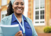 Practical steps towards teacher wellbeing A woman smiling while holding a folder, standing outdoors in front of a brick building with large windows.