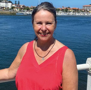 A woman in a red sleeveless top stands by a waterfront with buildings and boats in the background.