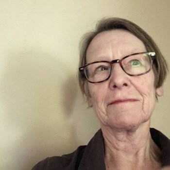 A person with short hair and glasses, wearing a dark top, looking slightly to the side against a plain wall.