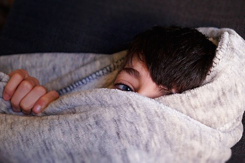 A child partially covered with a grey blanket, with only one eye and a hand visible, peeking out.