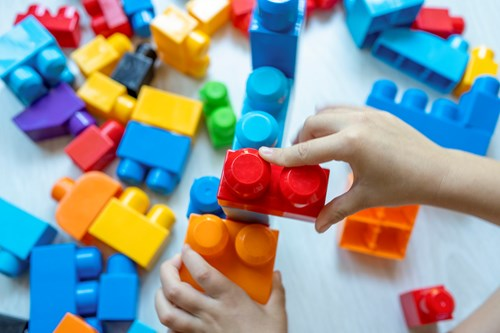 Child's hands holding interlocking red and orange plastic building blocks, with colorful blocks scattered on a light surface.