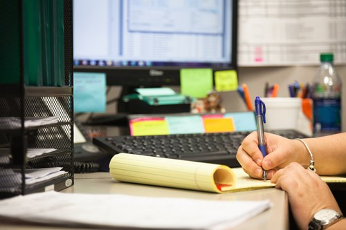 A person writing on a yellow notepad on a desk cluttered with office supplies, including a computer, keyboard, and sticky notes.
