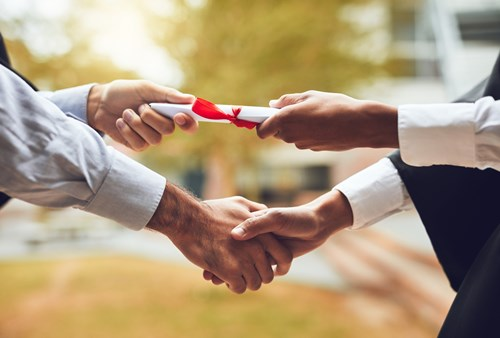 A person in a graduation gown shaking hands while receiving a diploma tied with a red ribbon.