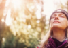 A feel-good lesson A woman wearing a beanie and jacket, with eyes closed, stands in a sunlit forest, suggesting a moment of relaxation and mindfulness.
