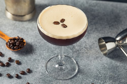 A creamy espresso martini garnished with coffee beans on a grey surface, surrounded by scattered coffee beans and bartending tools.