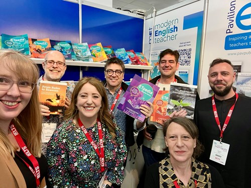 A group of people smiling at a book fair booth, holding up various educational books related to English teaching. The booth is decorated with book displays and has banners with 