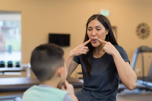 A woman demonstrating mouth movements to a young boy in a therapy or educational setting, likely for speech practice.