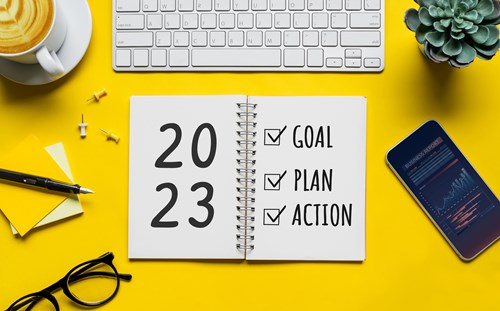 A desk workspace featuring a notebook open to a page with '2023' and a checklist reading 'Goal', 'Plan', 'Action'. Nearby are a keyboard, smartphone, coffee cup, eyeglasses, and a small plant, all set on a yellow background.