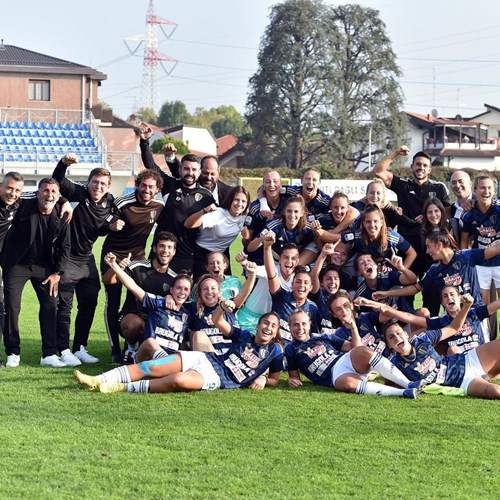 A women's football team and their staff celebrate together on a grassy field, wearing team uniforms with dark blue tops and white shorts. They are smiling, posing with arms raised in excitement, with a backdrop of a stadium and houses.