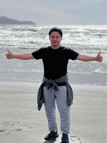 A person standing on a beach with arms outstretched and thumbs up, wearing a black t-shirt, grey sweatpants, and a jacket tied around the waist, with the ocean and hills in the background.