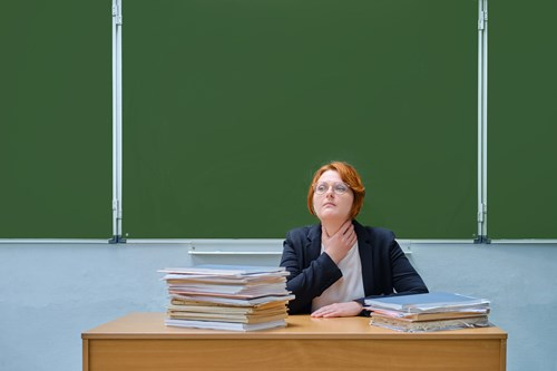 A person with short, reddish hair and glasses sits at a desk piled with papers, looking upward thoughtfully, against a backdrop of a green chalkboard.