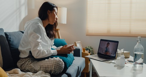A woman sitting on a sofa in cosy home attire, holding a mug and looking at a laptop screen showing a person, in a room with a table holding bottles and medications.