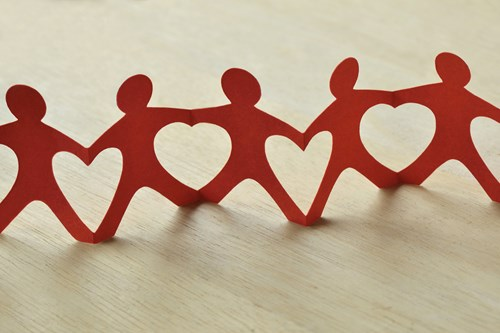A chain of red paper cutouts in the shape of people holding hands, each with a heart cutout in the center, placed on a light wooden surface.