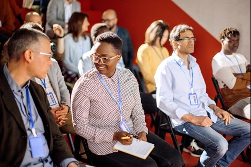 A diverse group of people seated in rows, some wearing conference badges. They appear engaged in conversation or listening intently, suggesting a seminar or workshop setting.