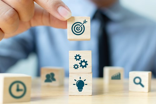 A person in a suit stacks wooden blocks with icons representing business concepts: target, gears and light bulb, and others like clock and chart in the background.