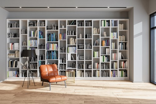 A modern living room with a large white bookshelf filled with books and decorative items against a light grey wall. There is a brown leather armchair and a tall black floor lamp on a wooden floor, with a small round glass side table nearby. Natural light enters through large windows on the right.