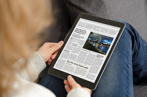 A person holds a tablet displaying a digital newspaper with various articles and images, including a photo of a cityscape at night.