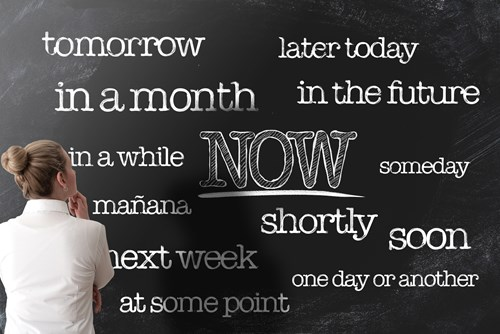 A person stands in front of a chalkboard with words like 'tomorrow,' 'later today,' 'in the future,' 'someday,' 'soon,' and 'NOW' written in different fonts and sizes.