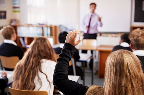 A classroom setting with students sitting at desks, one raising their hand, and a teacher at the front giving a lesson.