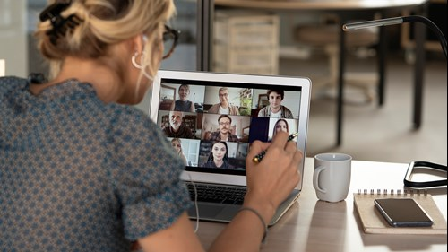 A person with blonde hair, wearing glasses and headphones, participates in a video conference on a laptop, showing multiple people on the screen. The person is holding a pen, with a notebook, smartphone, and coffee mug on the desk nearby.