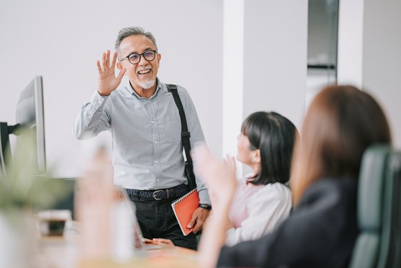 An elderly man with grey hair and glasses waves while holding a red notebook, standing beside an office desk with two women sitting and waving back.
