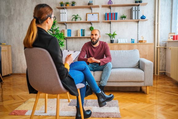 A man sitting on a sofa talking to a therapist who is taking notes on a clipboard, in a modern office with wooden floors and shelves decorated with plants and books.