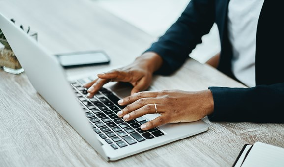 A person wearing a dark blazer is typing on a laptop at a wooden desk, with a smartphone and notebook nearby.