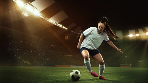 Female footballer in white jersey and navy shorts dribbling a soccer ball on a well-lit stadium field at night.